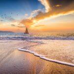 A Sailboat is Sailing Along the Ocean as Birds Fly Above and the Sun Sets on the Ocean Horizon