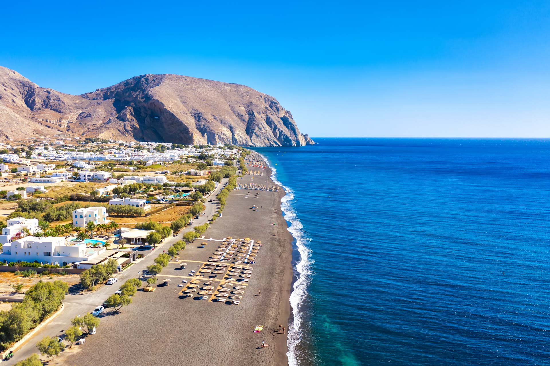 Top view aerial drone photo of black Perissa beach with beautiful turquoise water, sea waves and straw umbrellas. Vacation travel background. Aegean sea, Santorini Island, Greece.