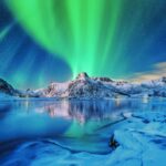 Aurora borealis over snowy mountains, frozen sea coast and reflection in water in Lofoten islands, Norway. Northern lights. Winter landscape with polar lights, ice in water. Starry sky with aurora